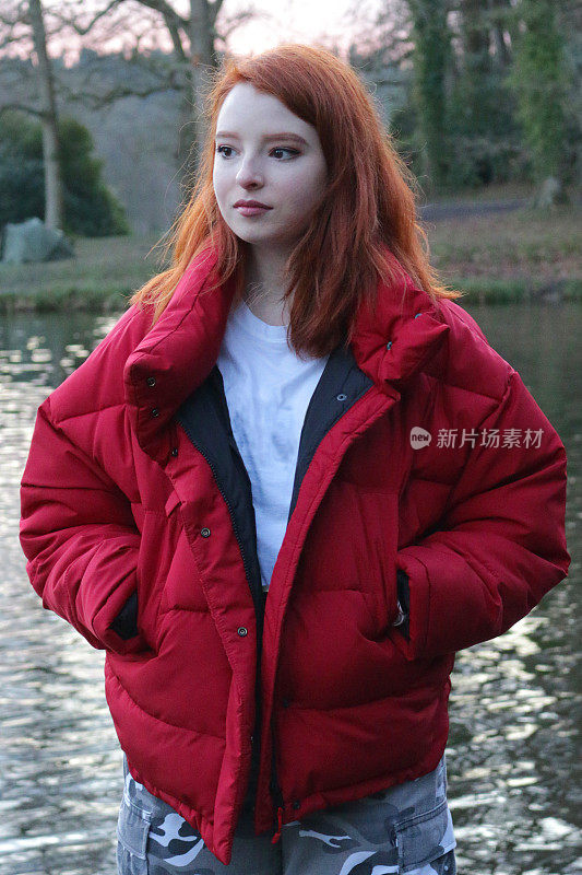 这是一个14 / 15岁的红发少女，皮肤苍白，脸上长着雀斑，穿着亮红色的羽绒服，外面罩着白色t恤和灰色迷彩长裤，站在户外的一个钓鱼湖旁边